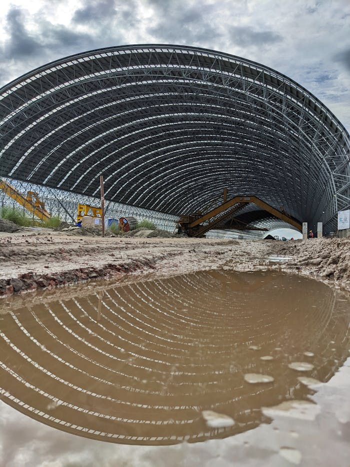 Puddle reflecting a large metal hangar roof at a construction site after rain.