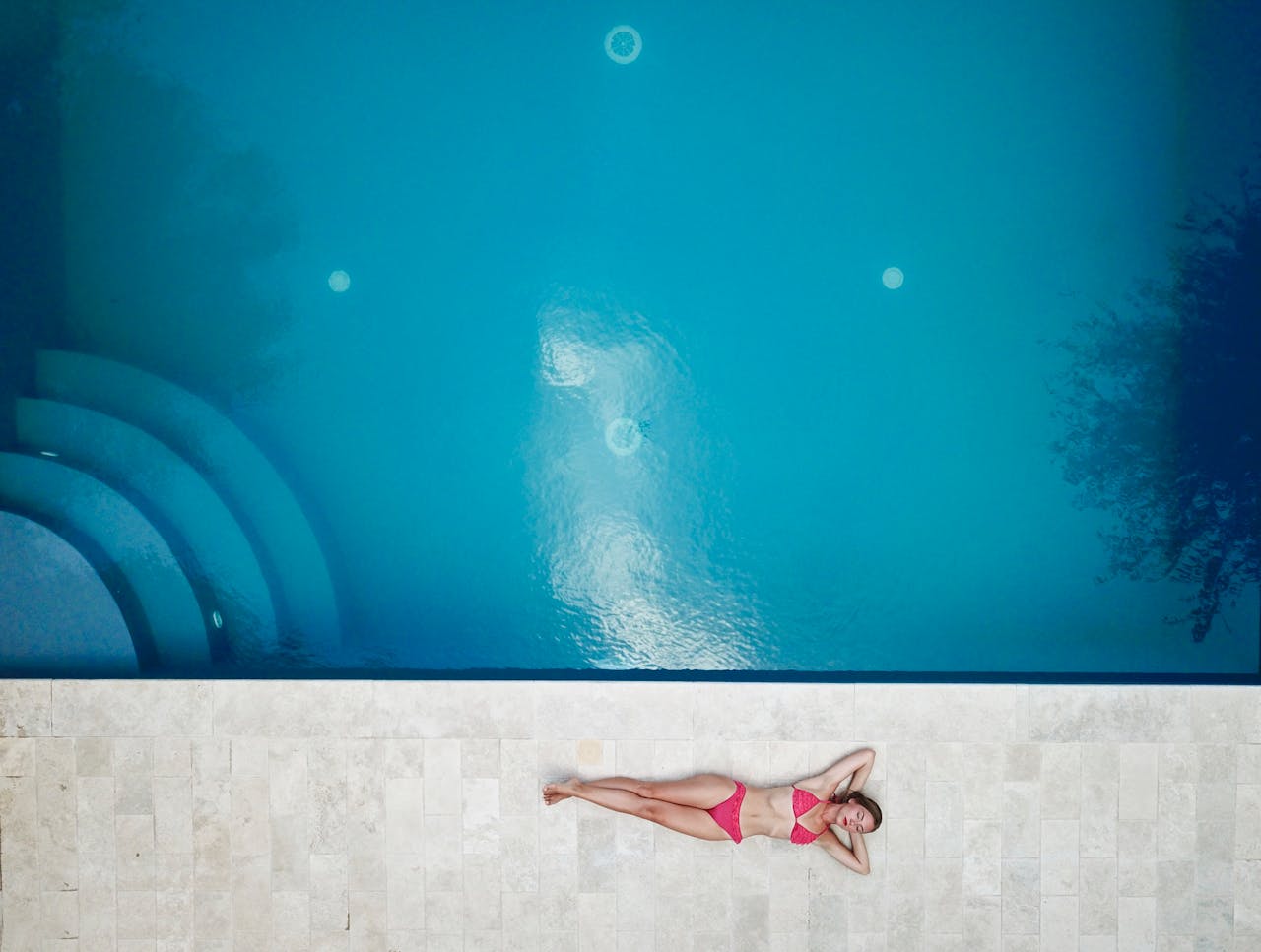 Bird's-eye view of a woman in a pink bikini relaxing by a bright blue swimming pool.